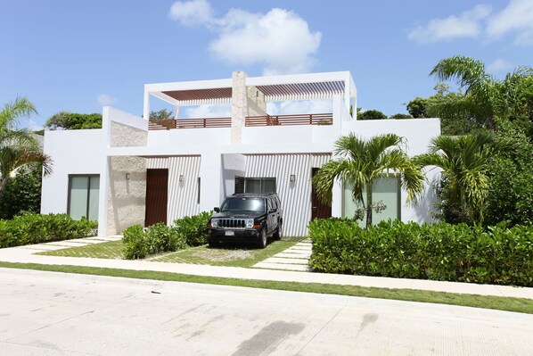 Una casa moderna de dos pisos con un coche en la entrada, rodeada de exuberante vegetación.