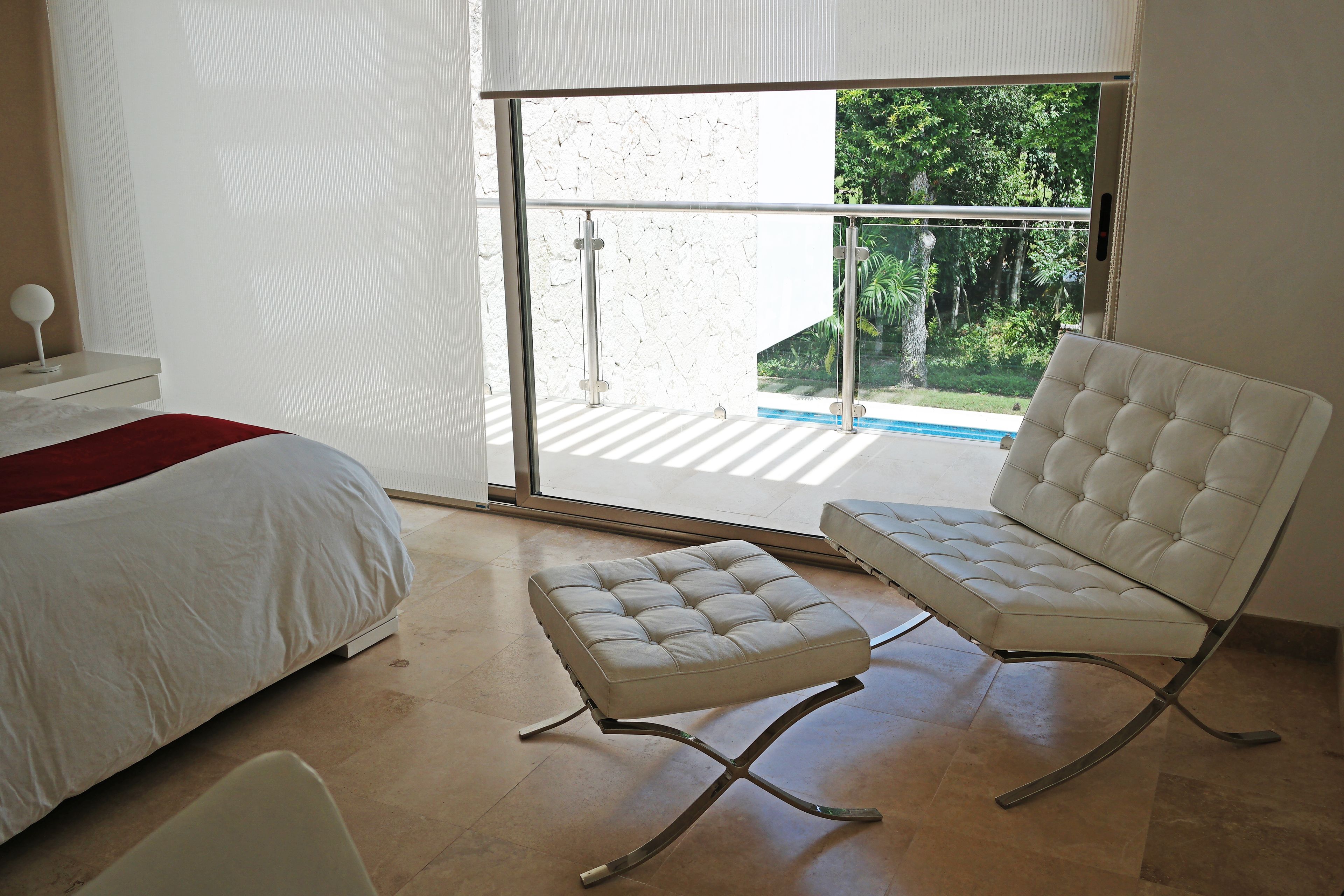 A modern bedroom with a bed, a white chair, and a glass door leading to a balcony with a view of greenery.