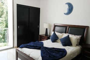 A bedroom with a bed, a nightstand, a lamp, a wardrobe, and a view of greenery outside.