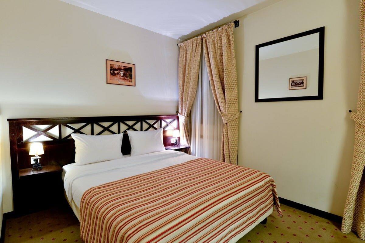 A hotel room with a bed, a nightstand, a lamp, a framed picture on the wall, and a window with curtains.