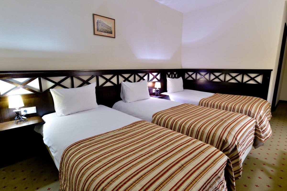 A hotel room with three beds, a wooden headboard, a framed picture on the wall, and a nightstand with a lamp.