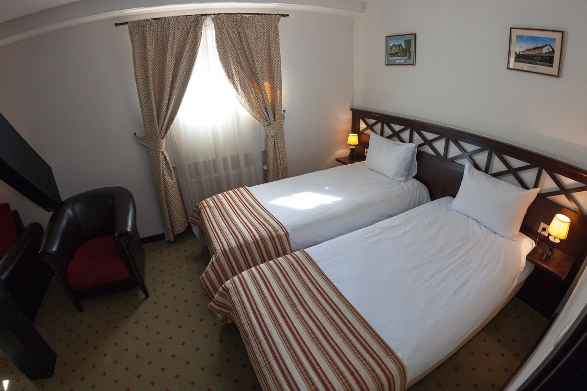 A hotel room with two beds, a chair, a window with curtains, and two wall-mounted lamps.