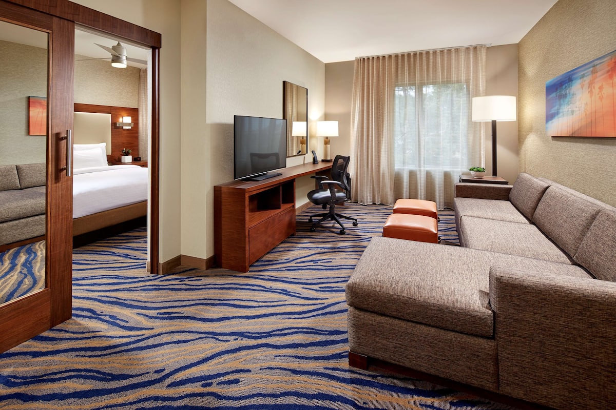 A hotel room with a bed, a TV, a desk with a chair, a sofa, and a large window with curtains.