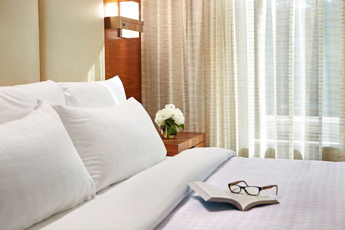 A neatly made bed with white linens, a bedside table with a vase of white flowers, and a book with glasses on it.