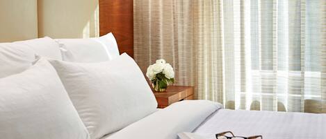 A neatly made bed with white linens, a bedside table with a vase of white flowers, and a book with glasses on it.