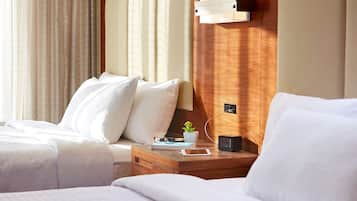 Habitación de hotel con dos camas, cabecero de madera, mesita de noche con una planta, vasos, un cuaderno y un reloj.