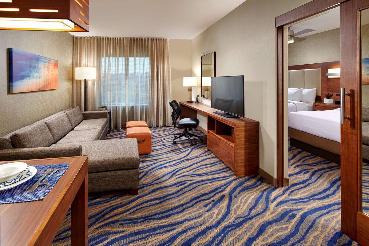 A hotel room with a sofa, a desk with a TV, a bed, and a large window with curtains.