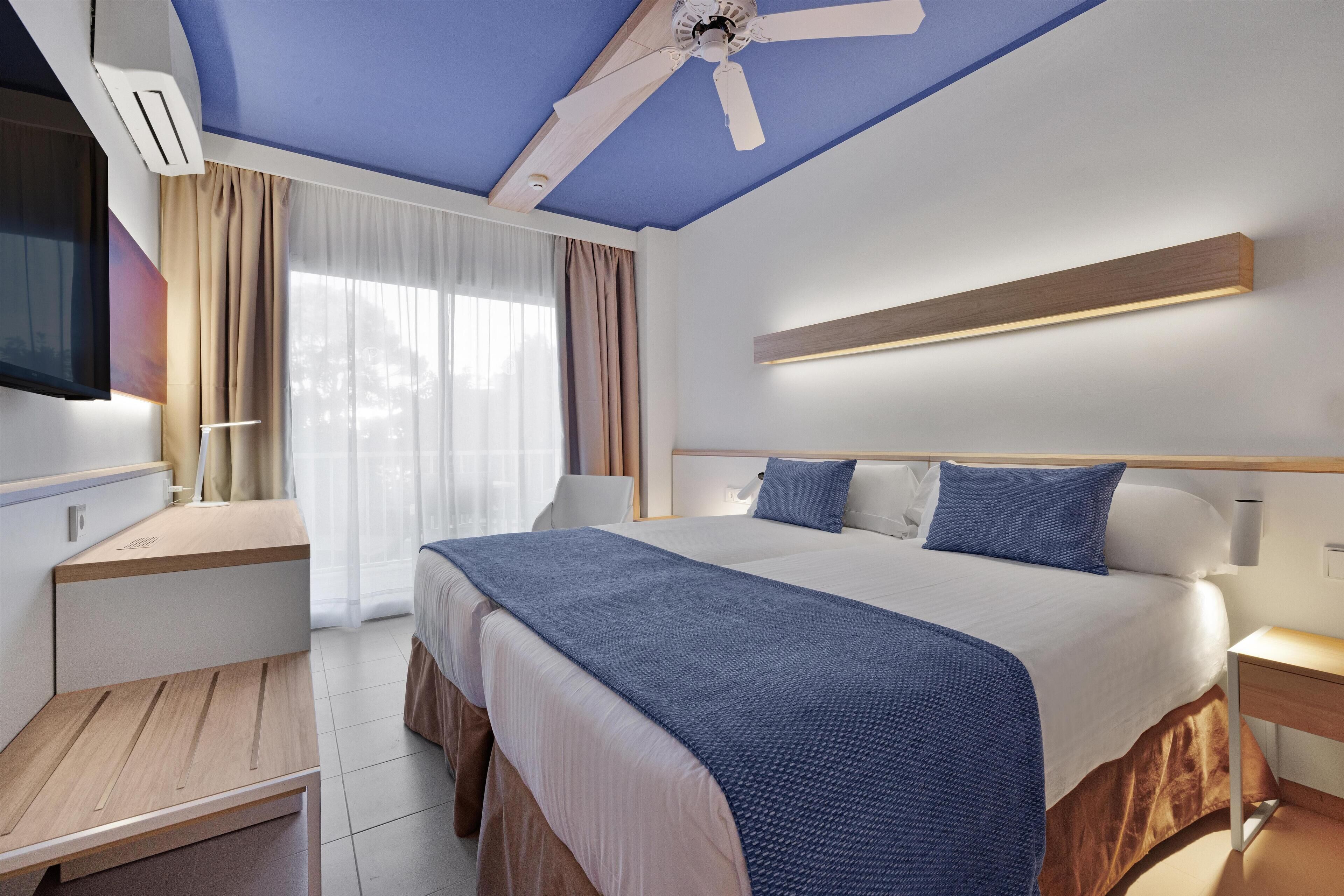 A modern hotel room with a large bed, a desk, and a ceiling fan.