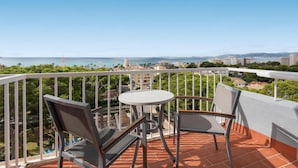Un balcón con mesa y sillas, con vistas a un paisaje urbano costero con edificios y zonas verdes.