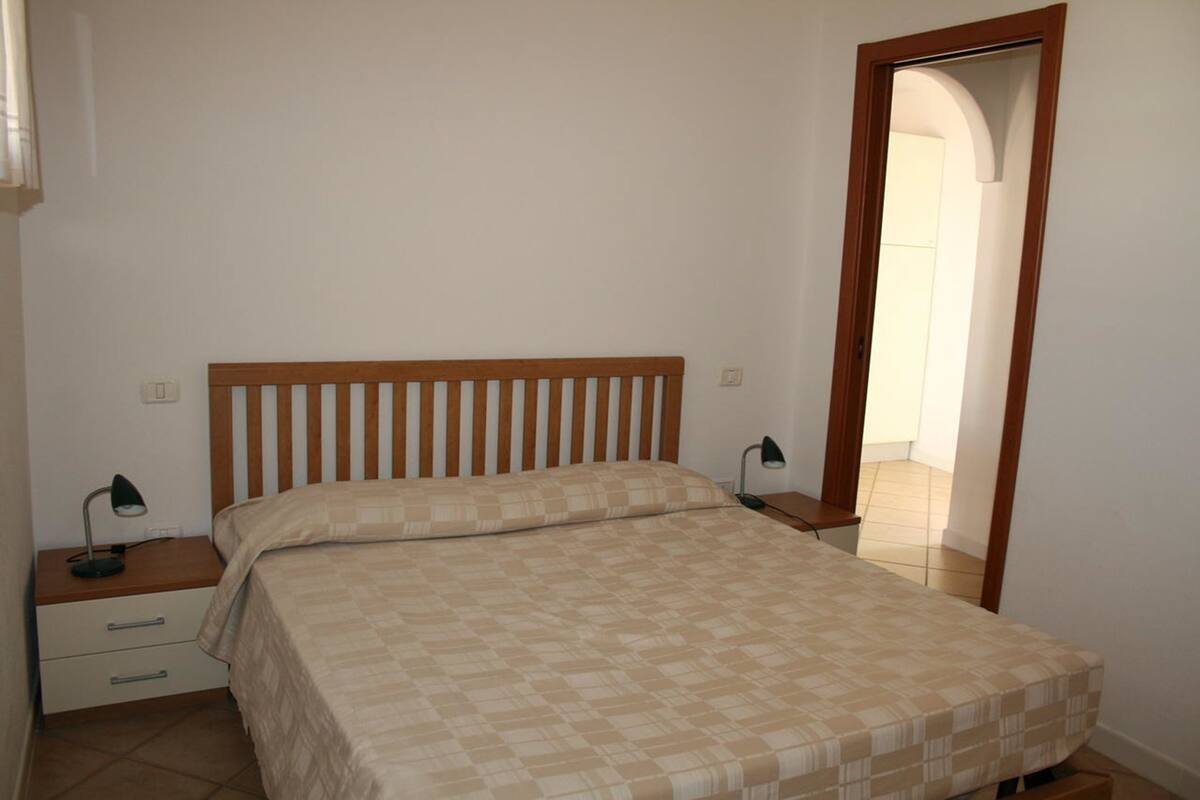 Une chambre à coucher avec un lit en bois, deux tables de chevet avec des lampes, une commode et une porte donnant sur une autre pièce.