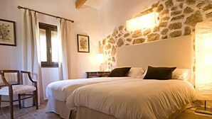A bedroom with two beds, a stone wall, a chair, and a window with curtains. - Vall de Pollença (Pollenca)