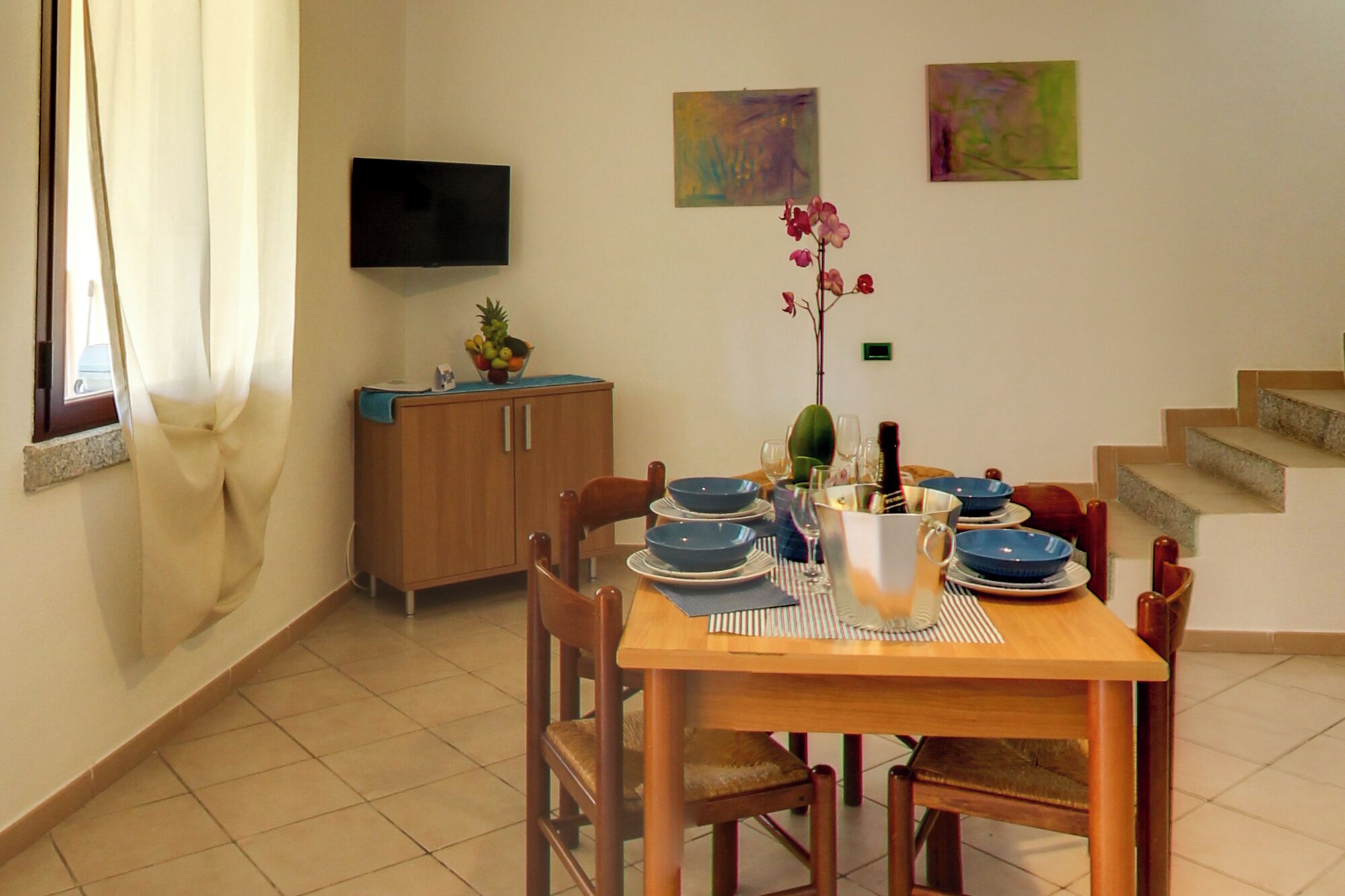 Un coin repas avec une table en bois, dressée pour quatre personnes, entourée de chaises. Un téléviseur est fixé au mur, au-dessus de la table.