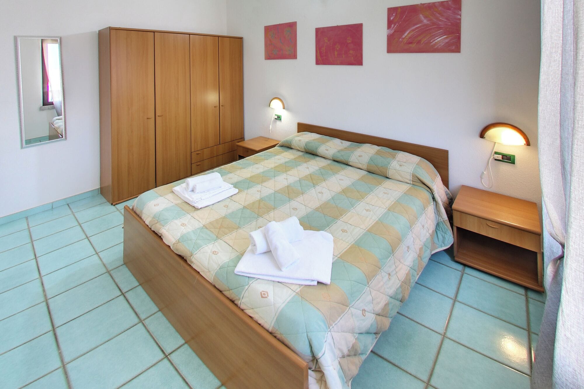 Ein ordentlich bezogenes Bett mit einer Decke, ein Holzschrank, Nachttische mit Lampen und ein gefliester Boden.