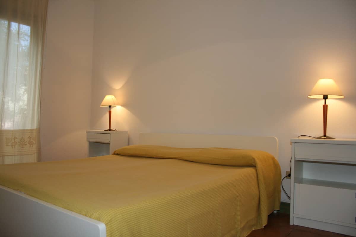 Ein Einzelbett mit einer gelben Decke, zwei Nachttische jeweils mit einer Lampe und ein Fenster mit Vorhängen.