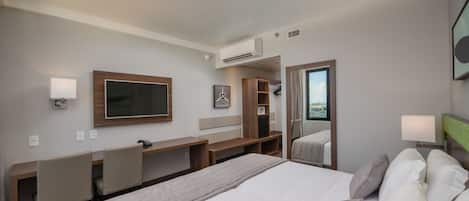 A hotel room with a large bed, a desk with a chair, a television, a mirror, and a window with a view.