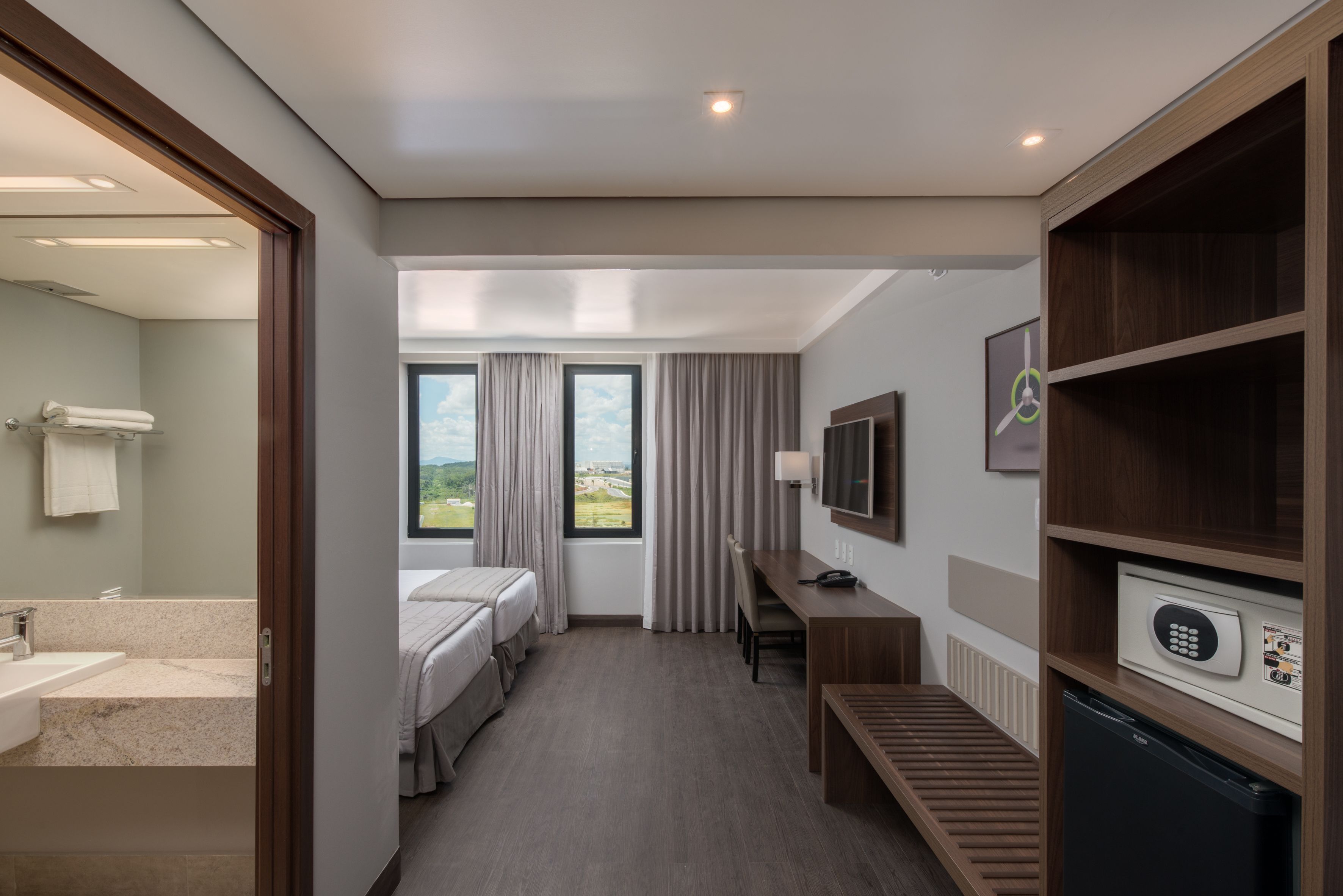 A hotel room with a bed, a desk, a TV, a minibar, and a bathroom with a sink and towel rack.