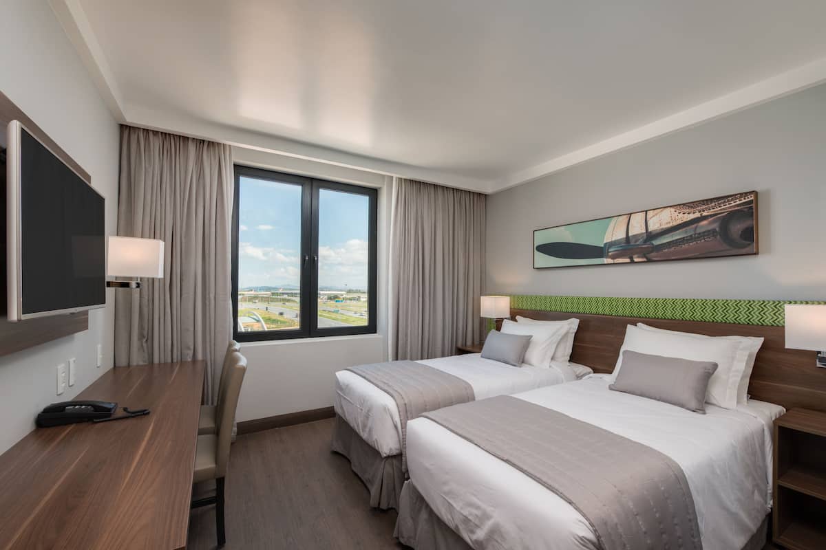 Habitación de hotel con dos camas, un escritorio, una silla, un televisor y una ventana con vistas al paisaje.
