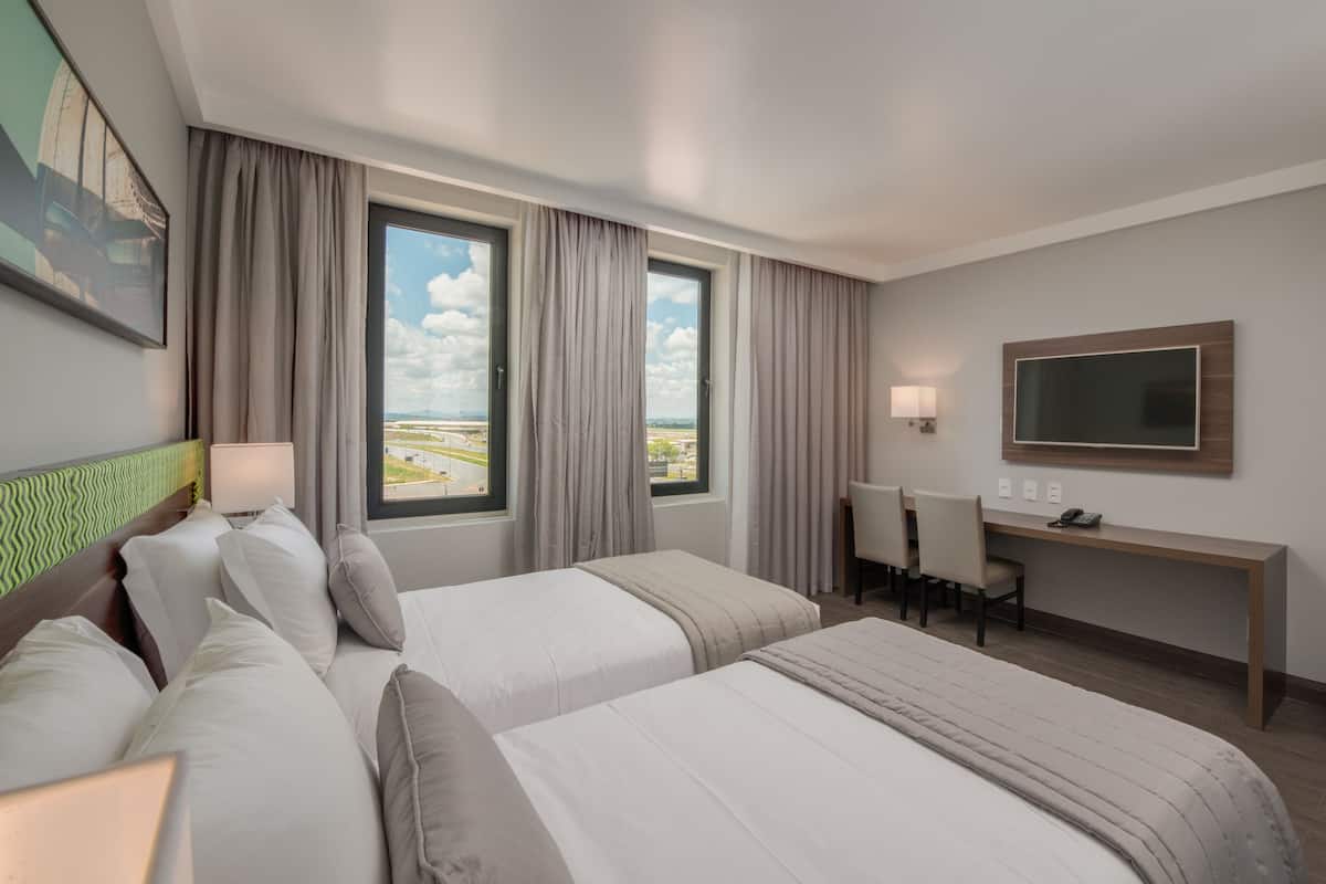 Habitación de hotel con dos camas, un escritorio, una silla, un televisor y ventana con vista a la ciudad.