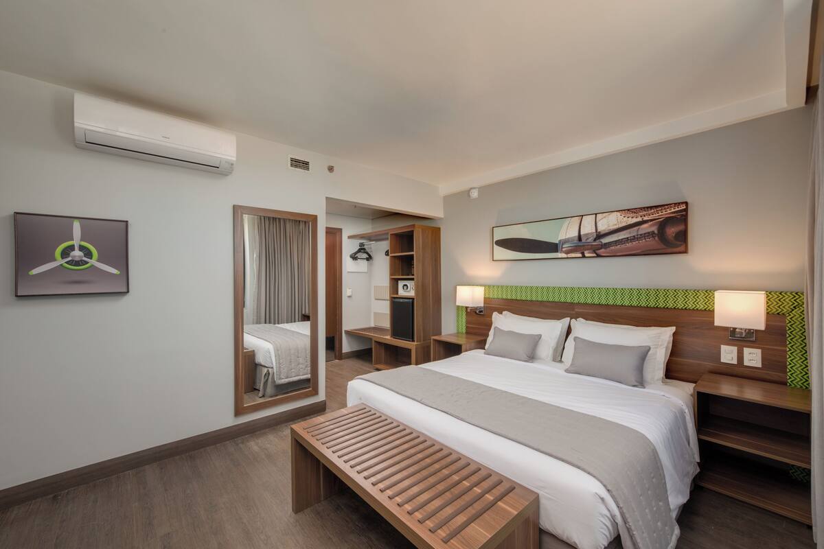 Un hotel moderno con una cama grande, mesitas de noche, un banco, un espejo y un cuadro en la pared.