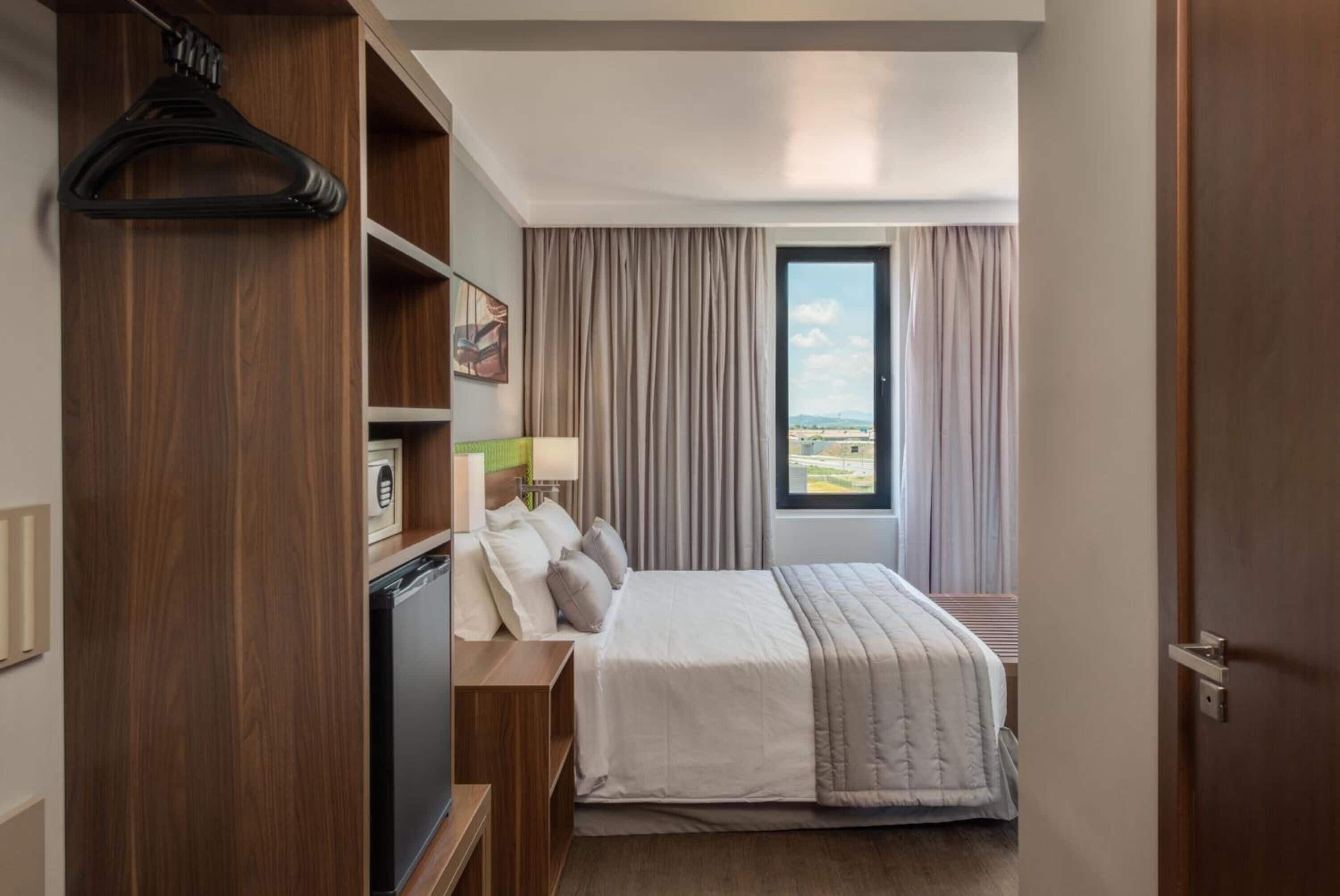 Quarto de hotel com uma cama, uma escrivaninha com abajur, um guarda-roupa e vista para o exterior.