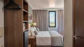 Quarto de hotel com uma cama, uma escrivaninha com abajur, um guarda-roupa e vista para o exterior.