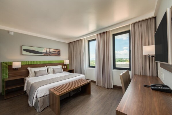 A hotel room with a bed, bedside tables, a desk, a chair, a TV, and a window with curtains.