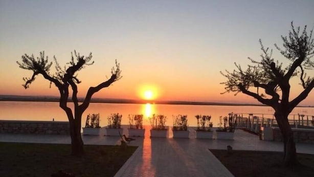 Vista al atardecer con un camino que lleva a un cuerpo de agua, bordeado por árboles y una barandilla.