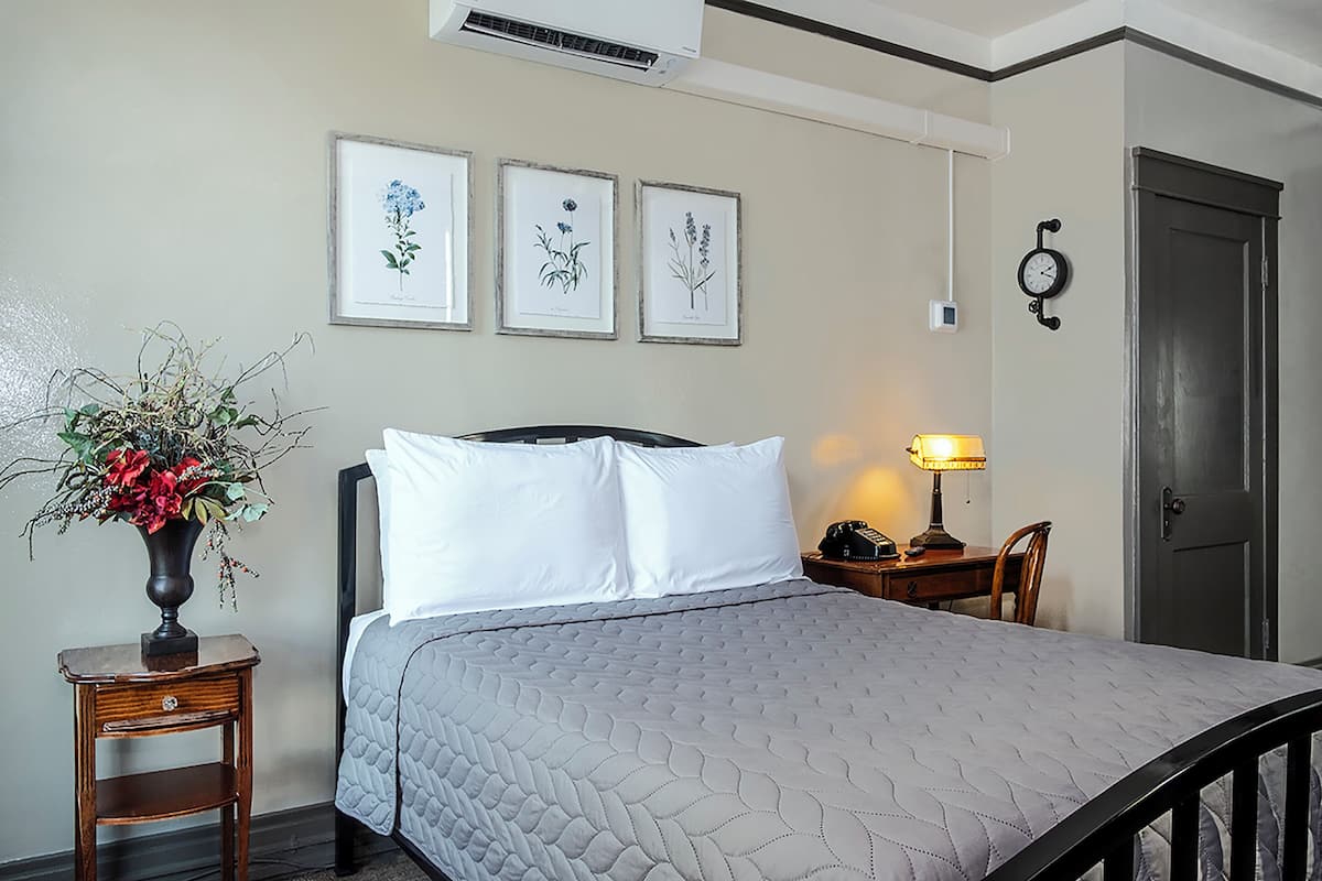 Una cama bien hecha con una colcha gris acolchada, una mesita de noche con una l ámpara y un jarrón con flores. Hay tres ilustraciones botánicas enmarcadas en la pared.