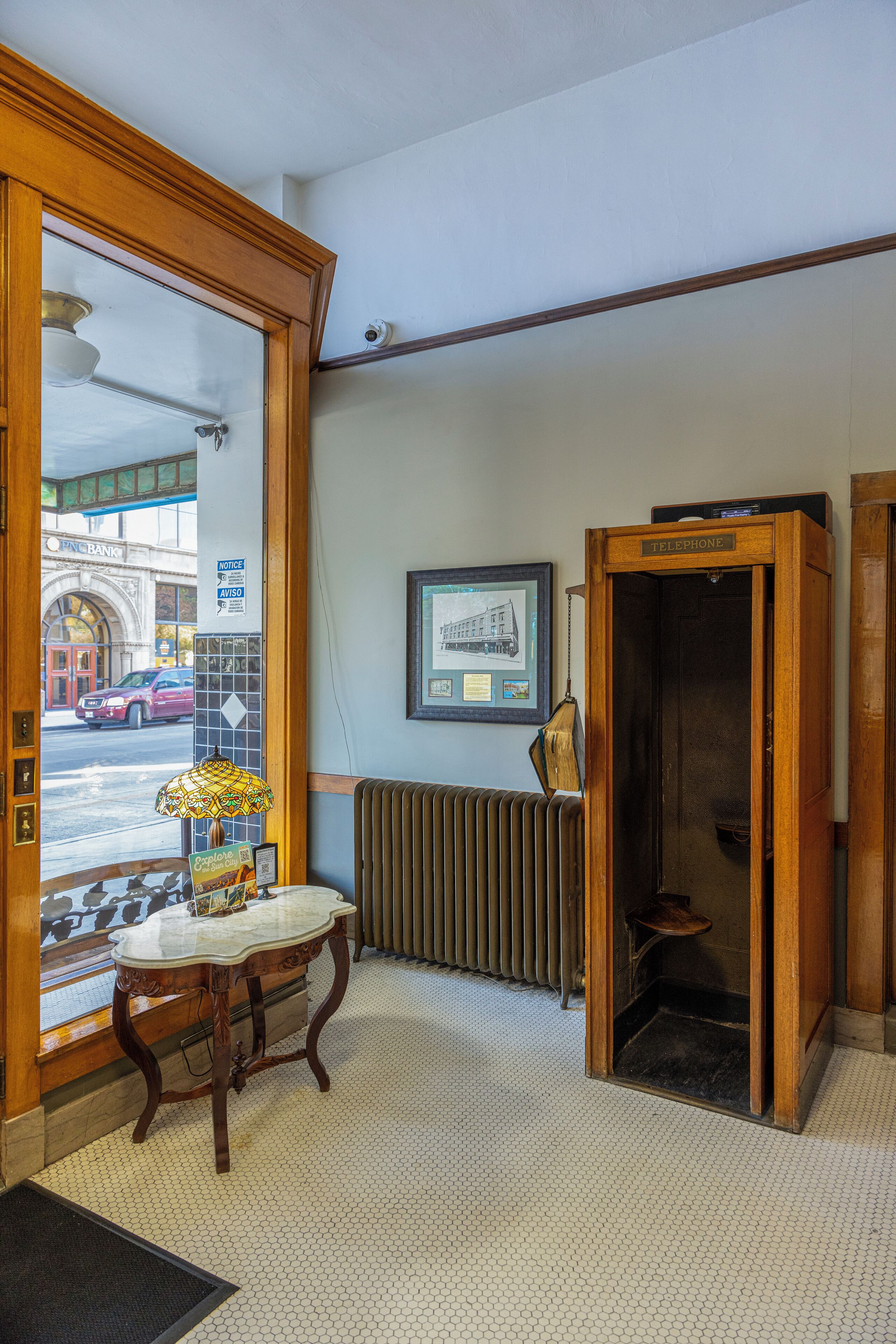 Vestíbulo de hotel con mostrador de recepción de madera, cabina telefónica, radiador y vista a la calle.