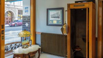 Vestíbulo de hotel con mostrador de recepción de madera, cabina telefónica, radiador y vista a la calle.