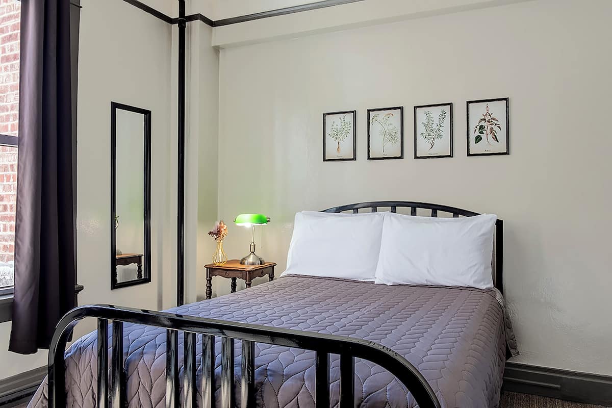Un dormitorio con cama, una mesita de noche con lámpara, un espejo y cuatro láminas botánicas enmarcadas en la pared.