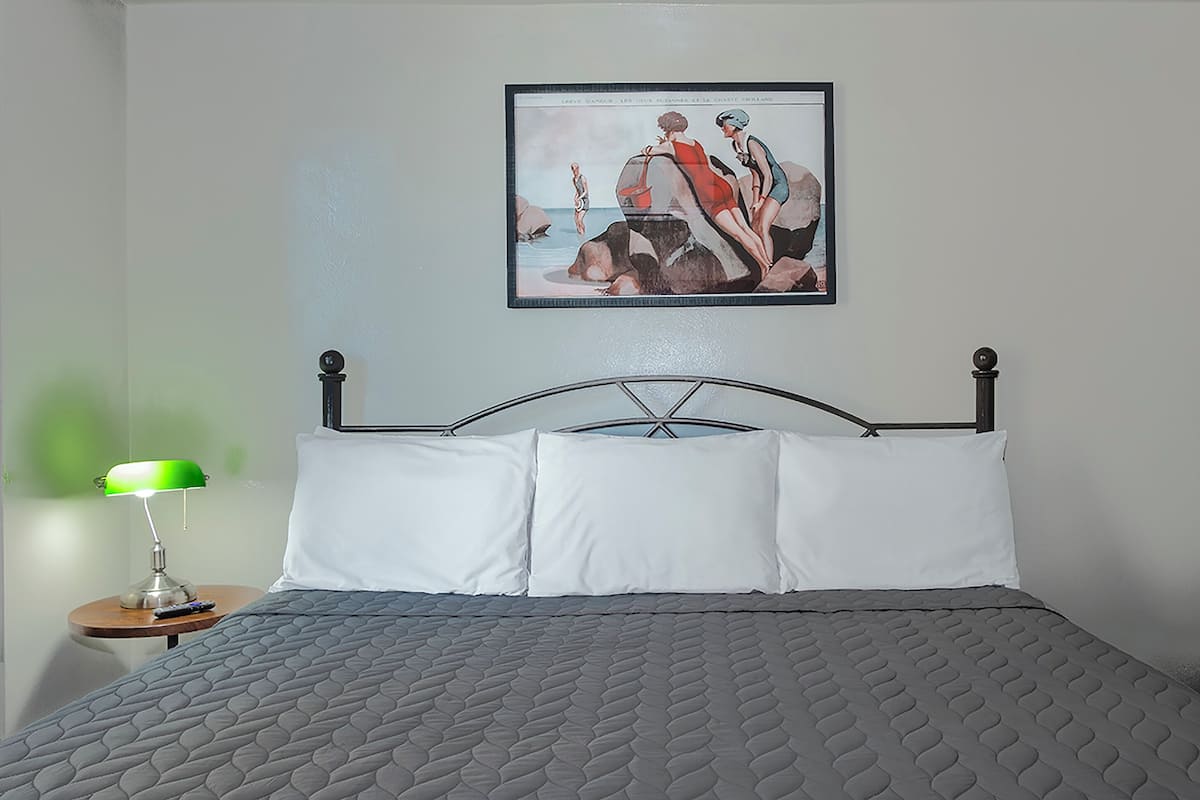 Una cama bien tendida con un edredón gris, una obra de arte enmarcada encima y una lámpara de escritorio verde sobre una mesita de noche.