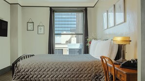 A hotel room with a large bed, a desk with a lamp, and a view of buildings through the window. - The Gardner  (El Paso)