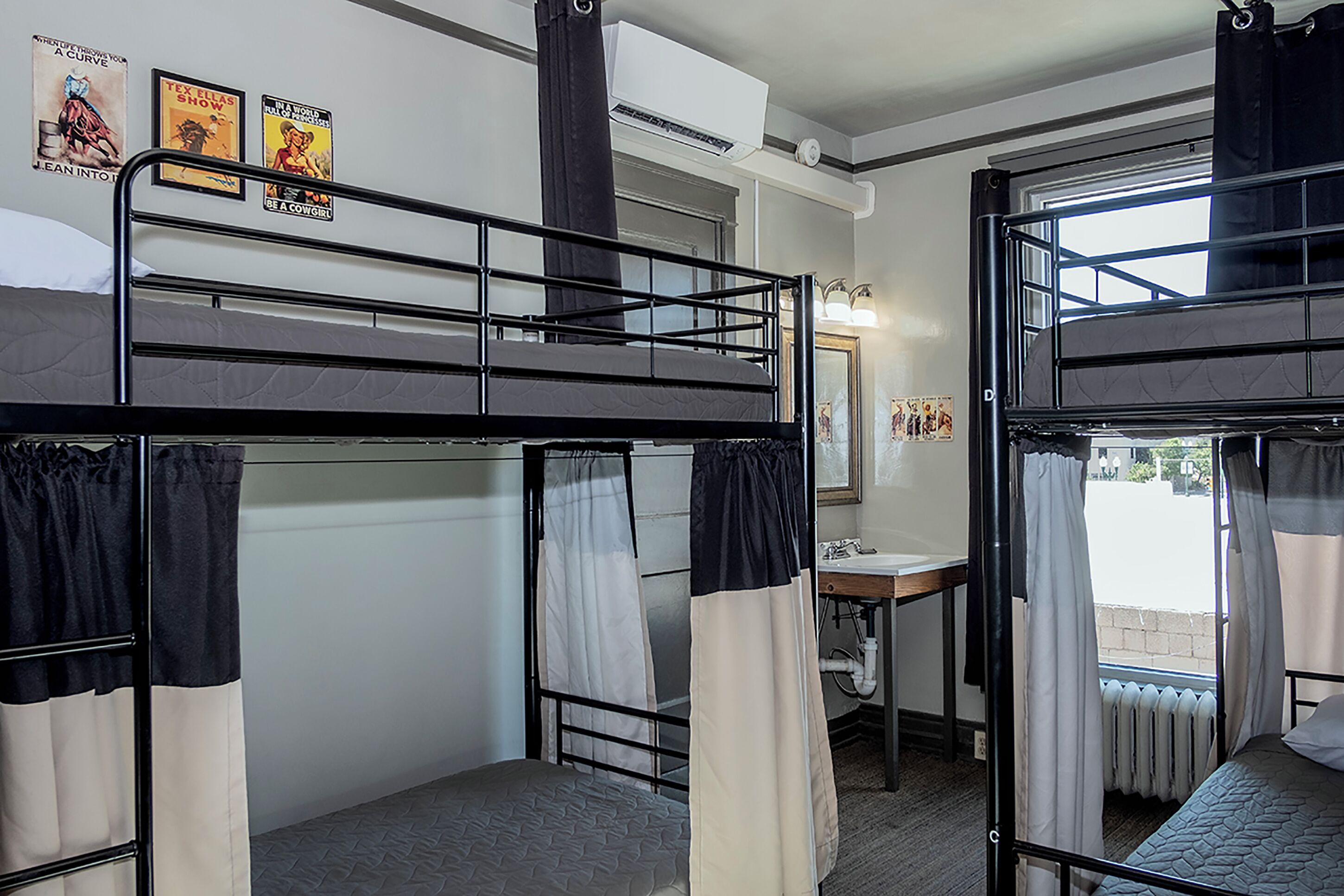 Quarto em dormitório com beliches, uma escrivaninha pequena com espelho e uma janela com cortinas.