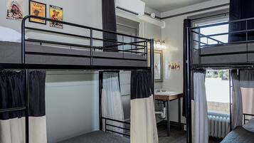 Quarto em dormitório com beliches, uma escrivaninha pequena com espelho e uma janela com cortinas.