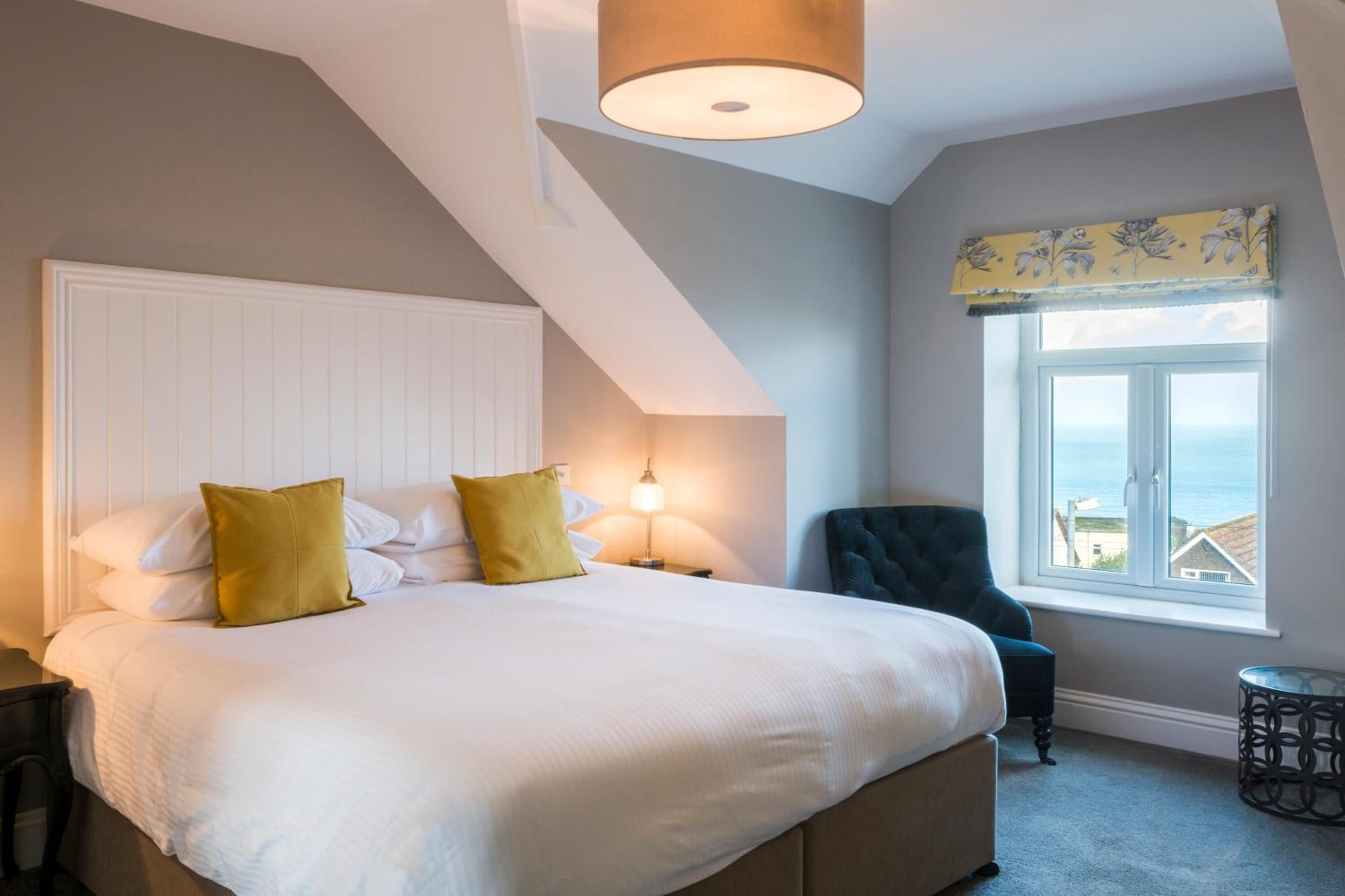 A bedroom with a large bed, a chair, a lamp, and a view of the sea through a window.