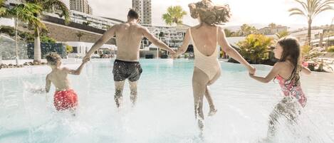 Een gezin van vier personen in een zwembad, met uitzicht op een hotel en palmbomen op de achtergrond.