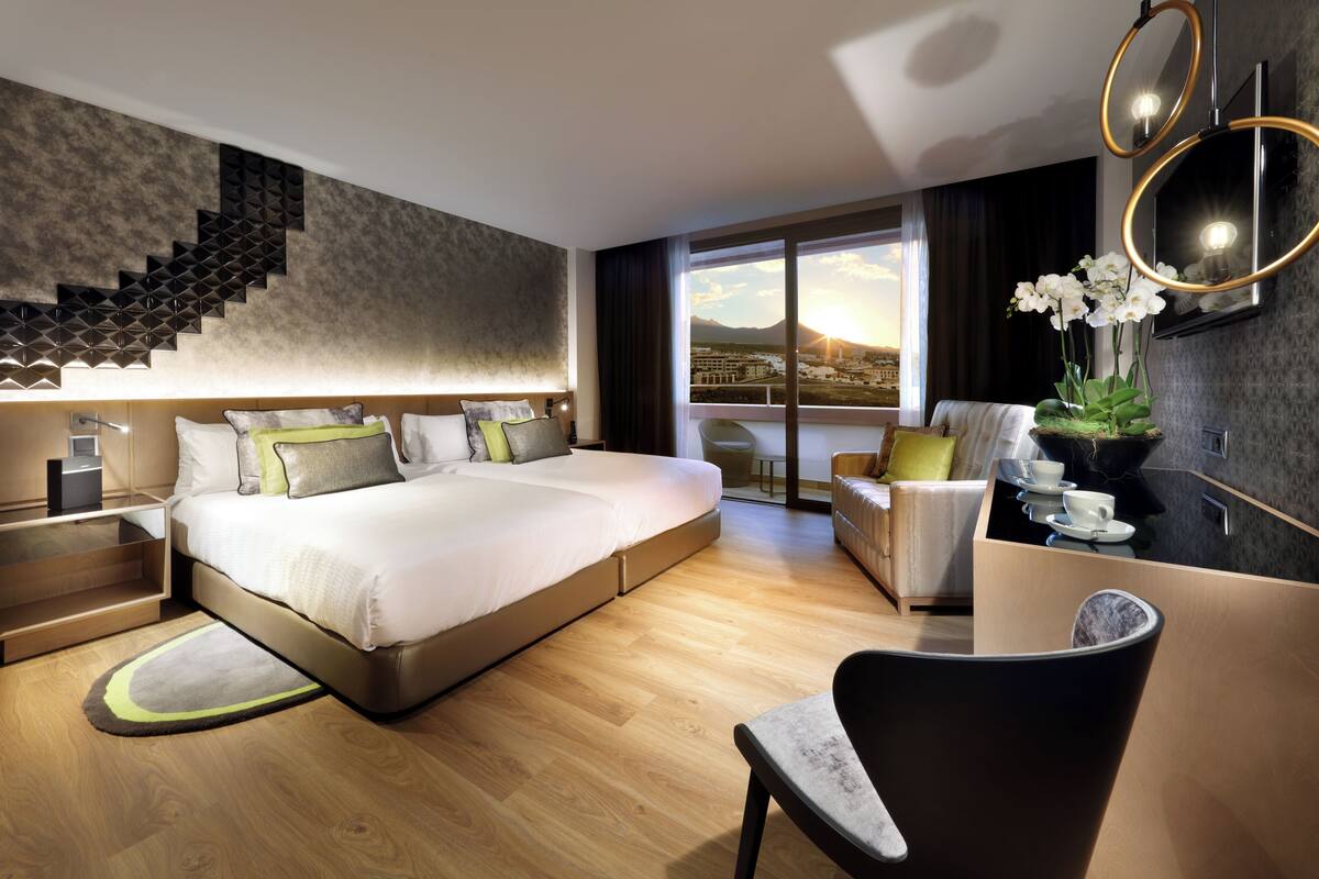 A modern hotel room with a large bed, a desk, a chair, a small table with a plant, and a view of mountains through the window.