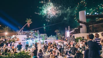 Un événement en plein air très fréquenté, avec des tables et des chaises, une scène et des feux d’artifice en arrière-plan.