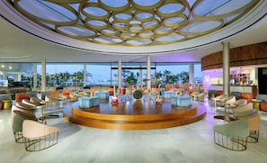 A spacious lounge area with a circular seating arrangement, a drum set, and a view of palm trees and a body of water. - Hard Rock Hotel Tenerife (Adeje)