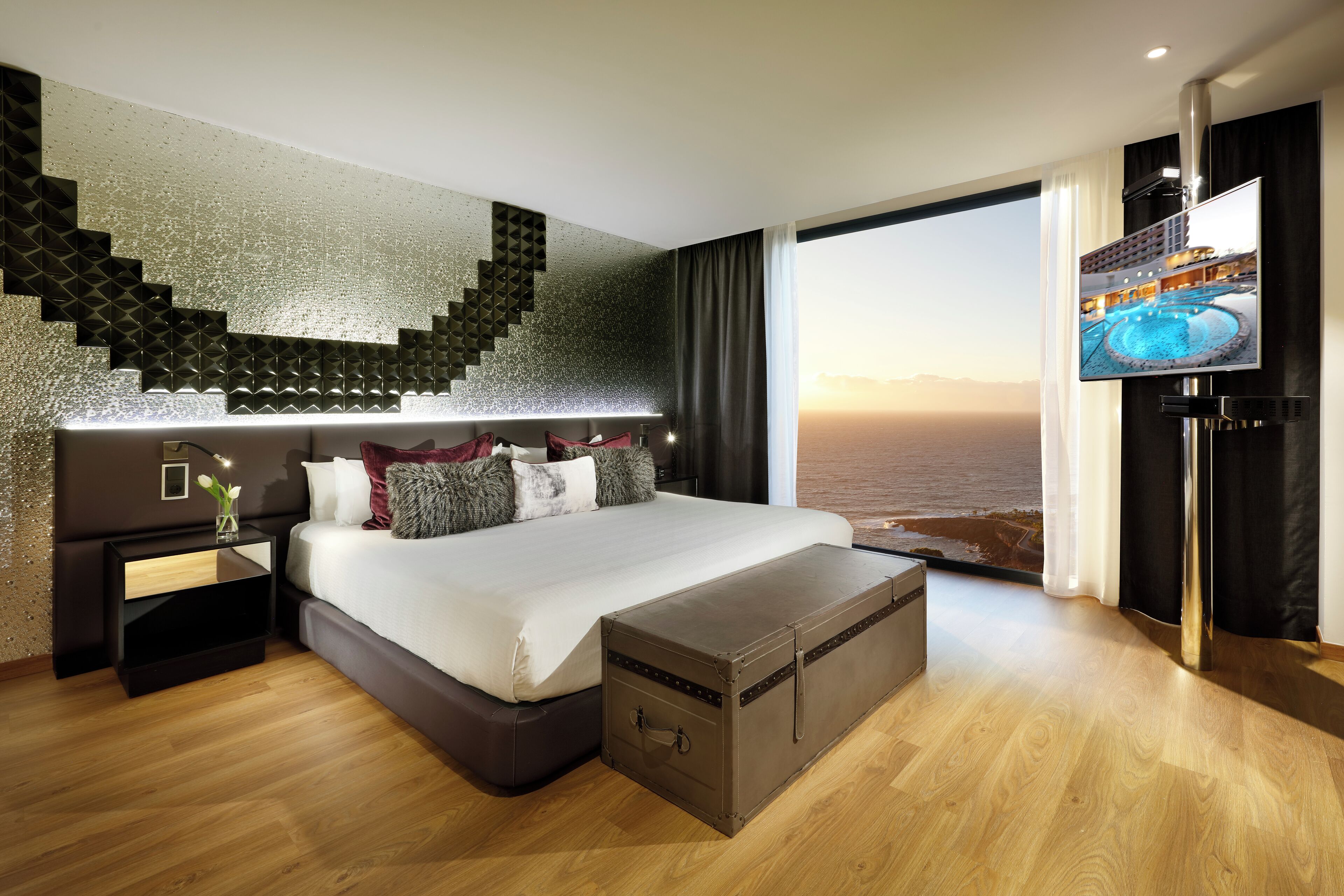 A modern hotel room with a large bed, a flat-screen TV, a wooden floor, and a view of the sea.
