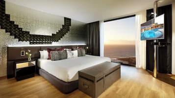 A modern hotel room with a large bed, a flat-screen TV, a wooden floor, and a view of the sea.