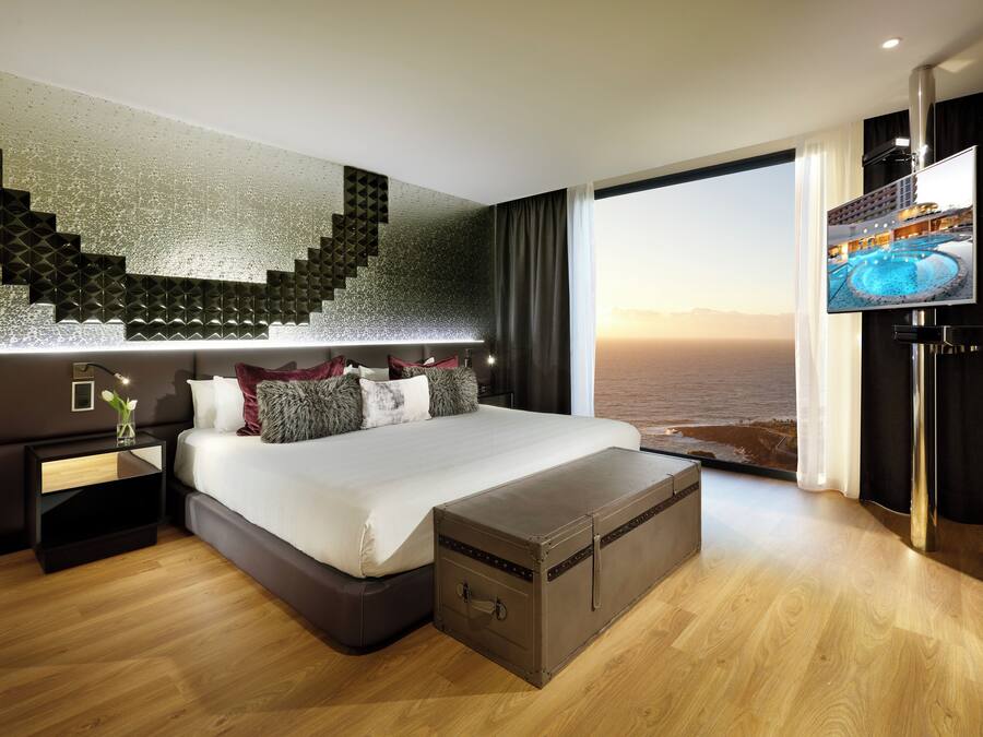 A modern hotel room with a large bed, a flat-screen TV, a wooden floor, and a view of the sea.