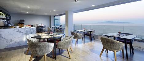 Uma zona de refeições moderna com vista para o mar, com bancadas de mármore, cadeiras contemporâneas e um bar com televisão.