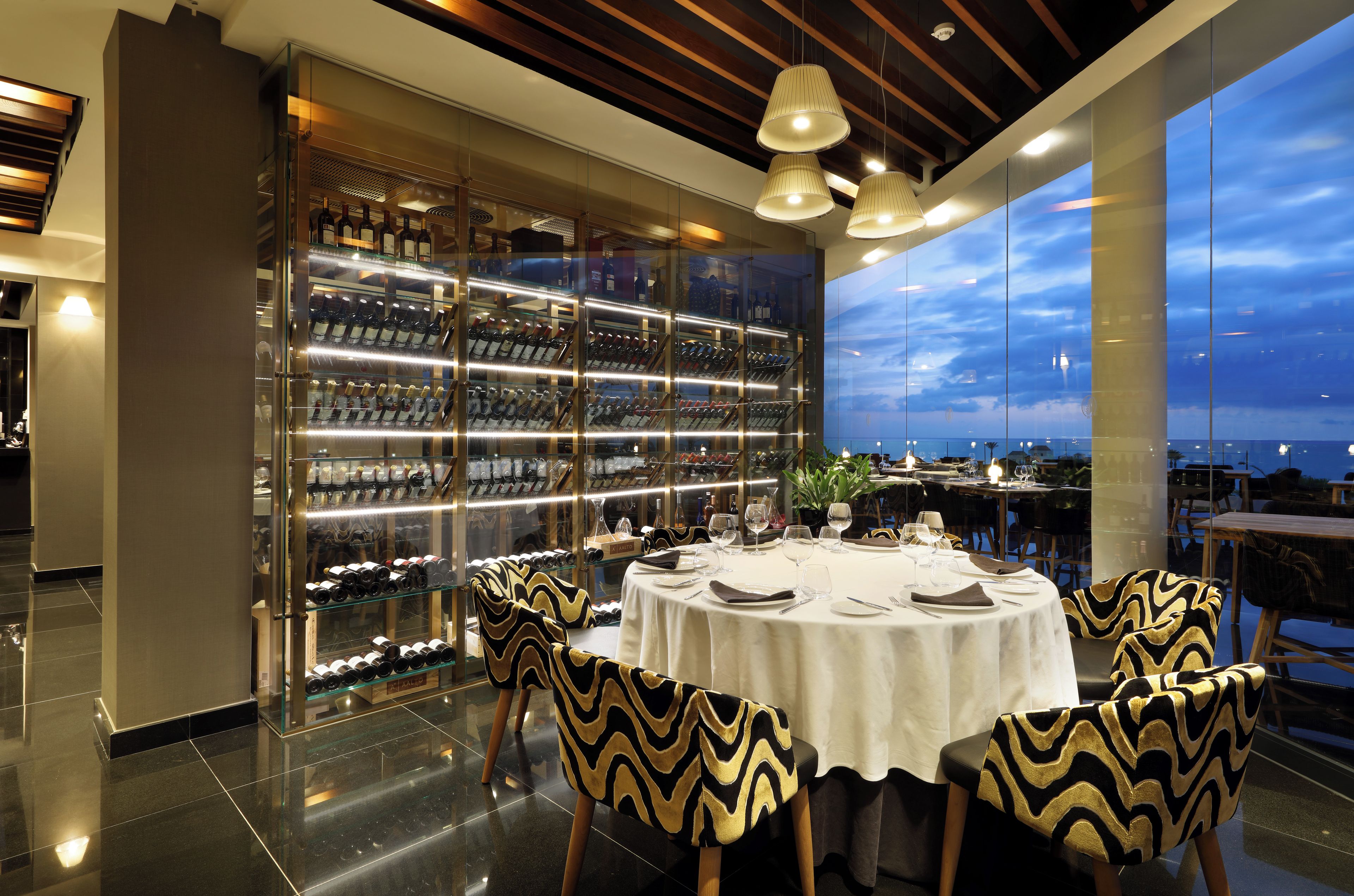 A modern dining area with a glass wine display, a round table set for dining, and a view of the sky through large windows.
