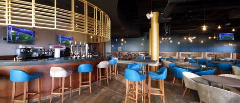 A modern bar area with wooden bar stools, blue chairs, and a large screen displaying sports.