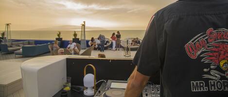 Un equipo de DJ con laptop, mezcladora y bebidas en una terraza con vista a una playa, con gente y sillas de descanso.
