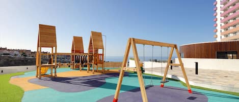 A playground with swings and a slide, surrounded by a building with balconies.