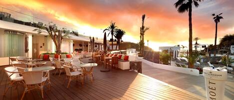 Eine Dachterrasse mit Korbmöbeln, Palmen und Blick auf den Sonnenuntergang.
