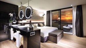 Habitación de hotel moderna con una cama grande, dos mesitas de noche con espejos redondos, un tocador con un espejo grande, vistas al atardecer por la ventana y una pequeña zona de estar con una silla.